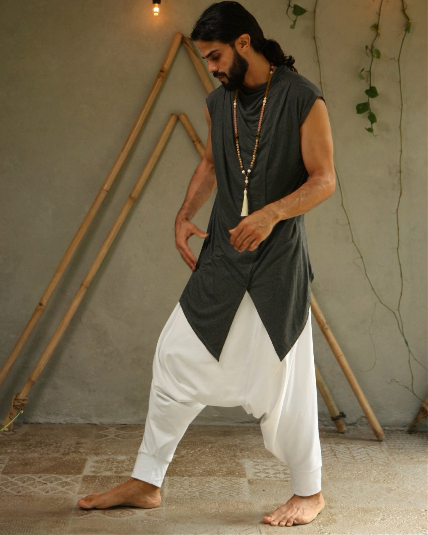 Man wearing glorka Jedi sleeveless dark shirt and white pants standing in a room with wooden frames and string lights.
