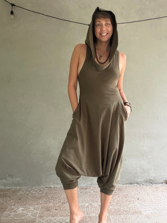 A woman wearing an olive green hooded jumpsuit and baggy harem-style legs, standing barefoot against a textured gray wall.