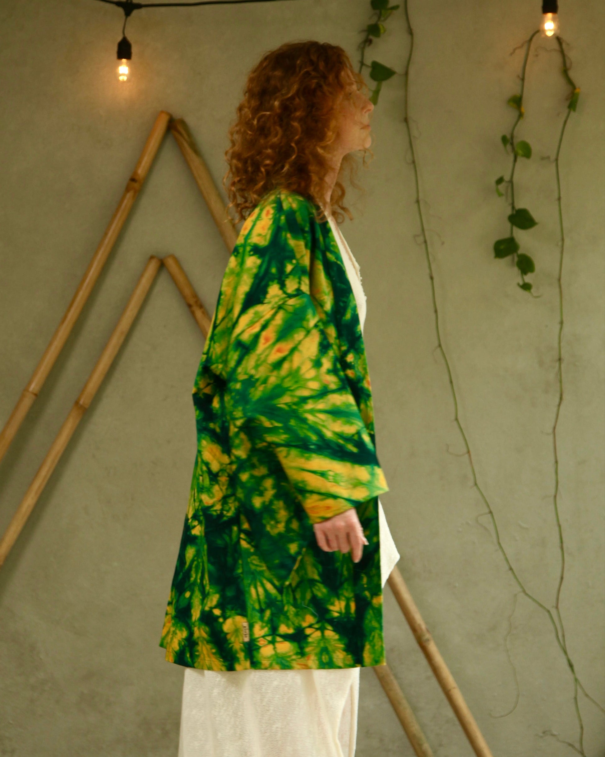 Person wearing a green and yellow tie-dye jacket in a room with wooden sticks and plants.