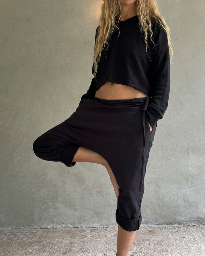 A woman with long blonde hair poses in a black cropped Glorka Pulse crop top and matching black low-waisted TOM pants against a textured gray concrete wall with hanging string lights.
