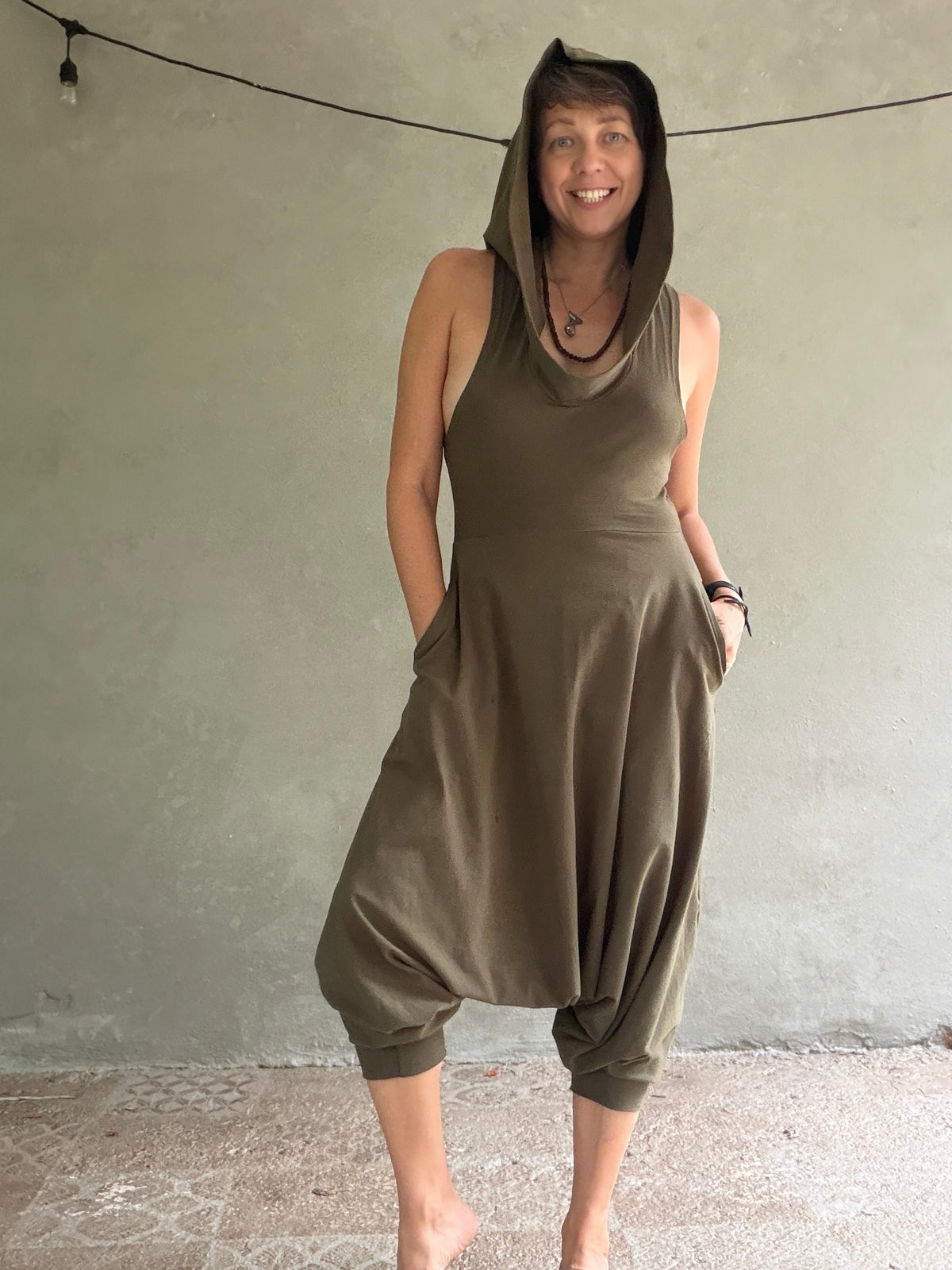 A woman wearing an olive green hooded jumpsuit and baggy harem-style legs, standing barefoot against a textured gray wall.