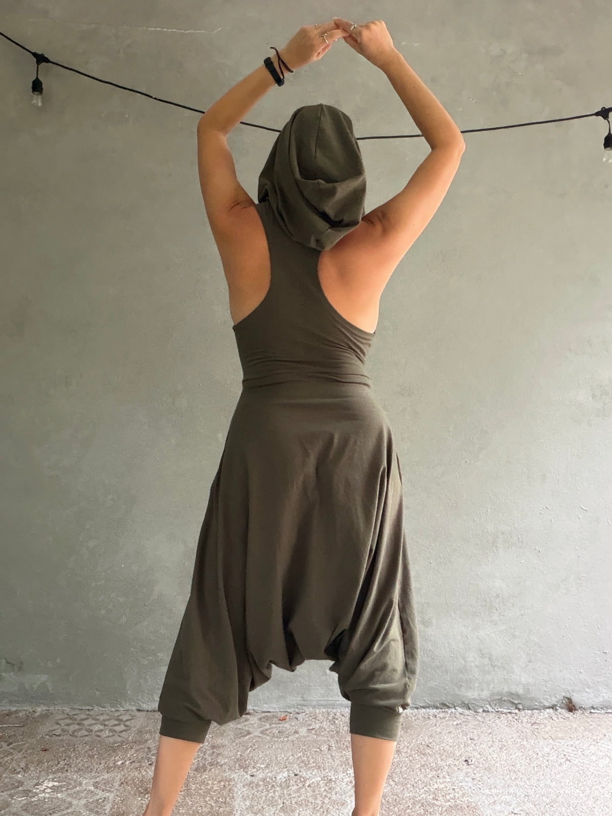 A woman wearing an olive green harem-style glorka jumper with a matching draped head covering, photographed from behind against a textured gray wall.