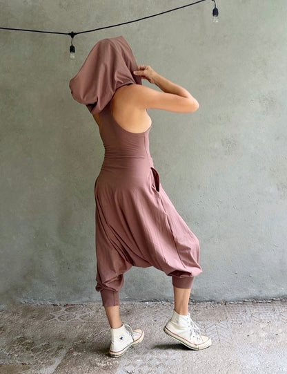 A person wearing a dusty rose-brown harem-style jumpsuit with a matching draped head covering, standing against a textured gray wall.