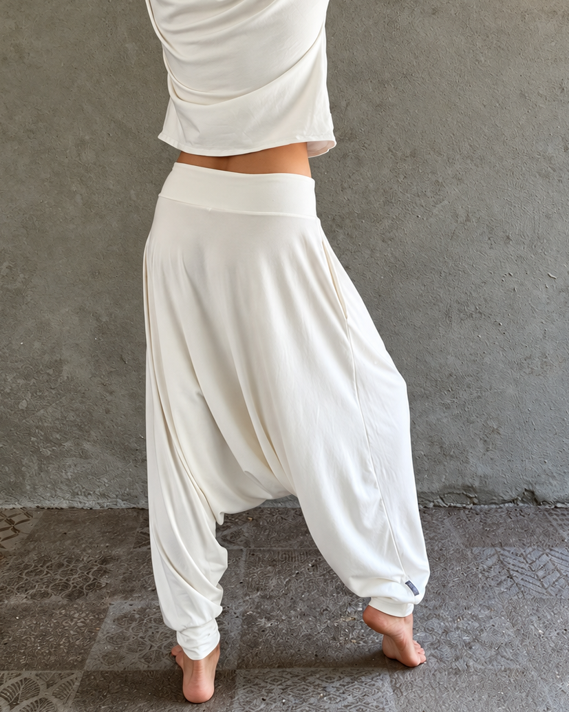 A woman stands barefoot against a textured gray wall, wqwhite harem-style glorka pants and a straw hat with black and white stripes. Warm string lights hang above, and the floor is a rustic stone surface.earing 