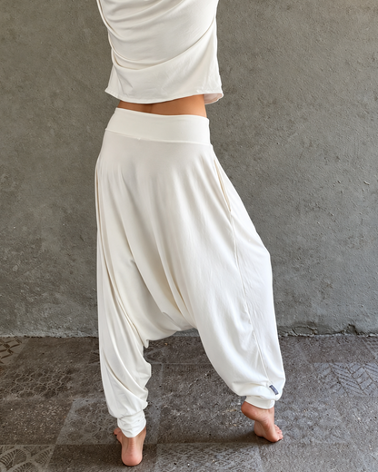 A woman stands barefoot against a textured gray wall, wqwhite harem-style glorka pants and a straw hat with black and white stripes. Warm string lights hang above, and the floor is a rustic stone surface.earing 