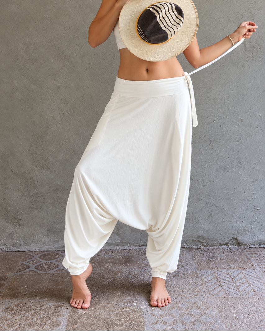 A woman stands barefoot against a textured gray wall, holding a large white harem-style glorka pants and a straw hat with black and white stripes. Warm string lights hang above, and the floor is a rustic stone surface.