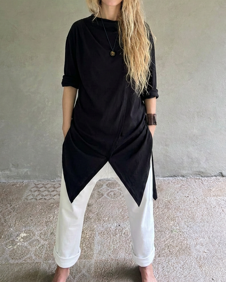A woman stands barefoot against a textured gray wall, wearing Glorka black asymmetrical tunic top with white pants.