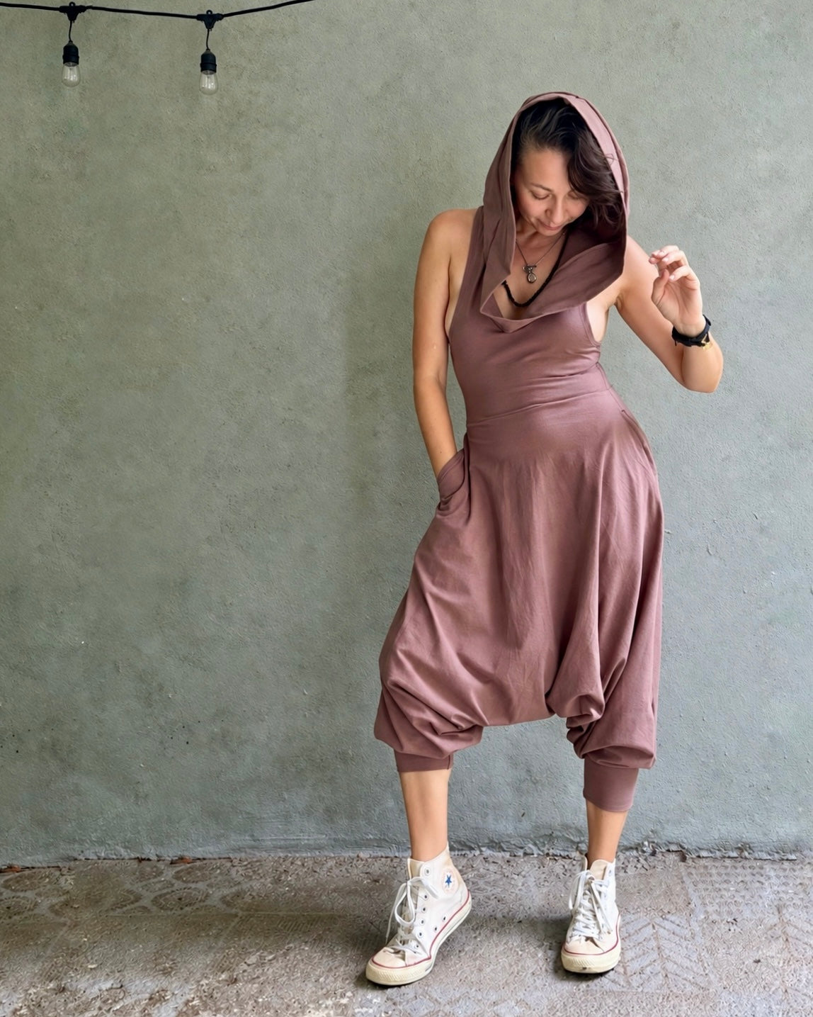 A woman wearing a dusty rose sleeveless hooded glorka jumpsuit with baggy harem-style legs and white sneakers, posing against a textured gray wall.