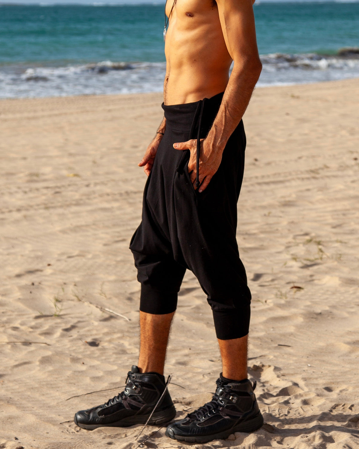 A shirtless man stands on a sandy beach wearing loose black pants and black boots, with the ocean and blue sky in the background.