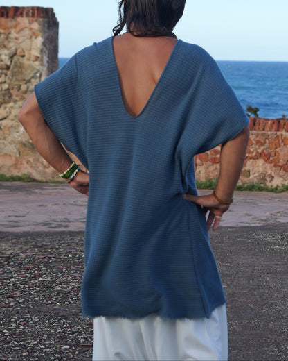 Person wearing a blue open-back top and white pants standing by a coastal wall.