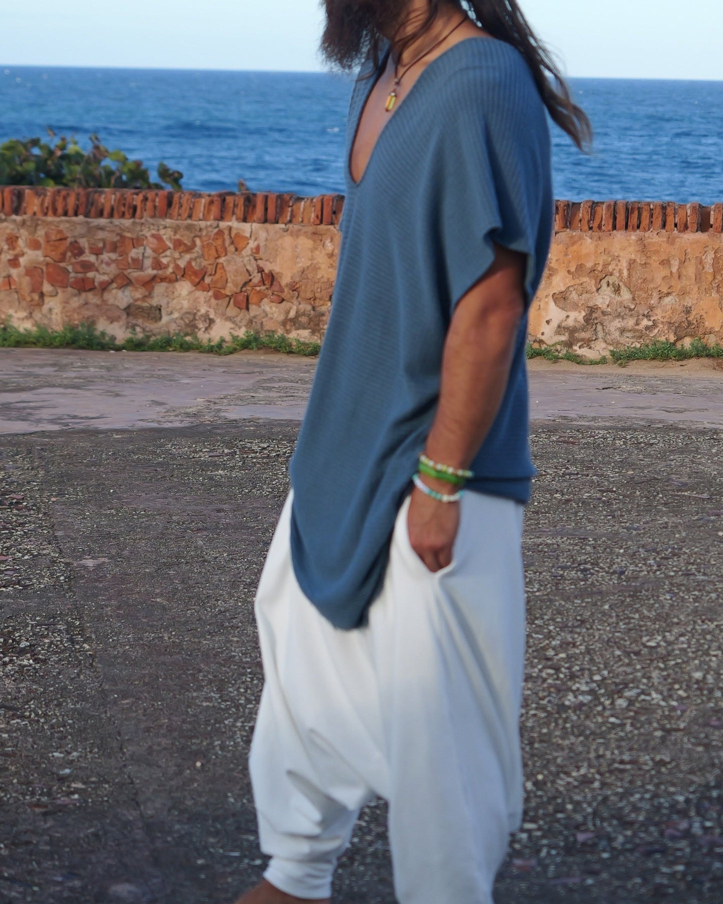Man with long hair and beard wearing a blue shirt and white pants standing on a road with ocean in the background.