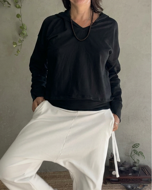 A woman posing in a black long-sleeve glorka Pulse crop top and off-white relaxed-fit glorka TOM pants against a textured gray wall and woven rug.