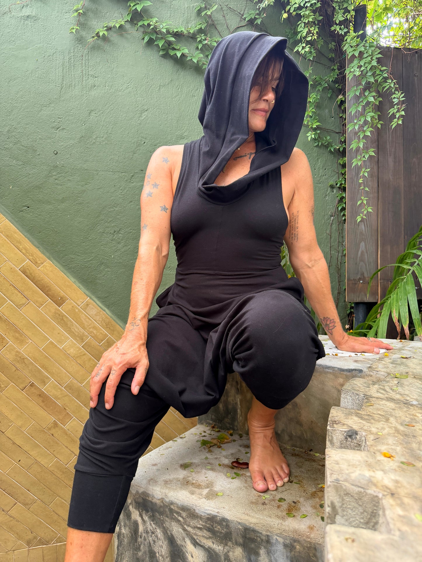 A woman wearing a black hooded sleeveless Glorka Aura jumpsuit crouches on stone steps outdoors against a green wall with climbing vines.