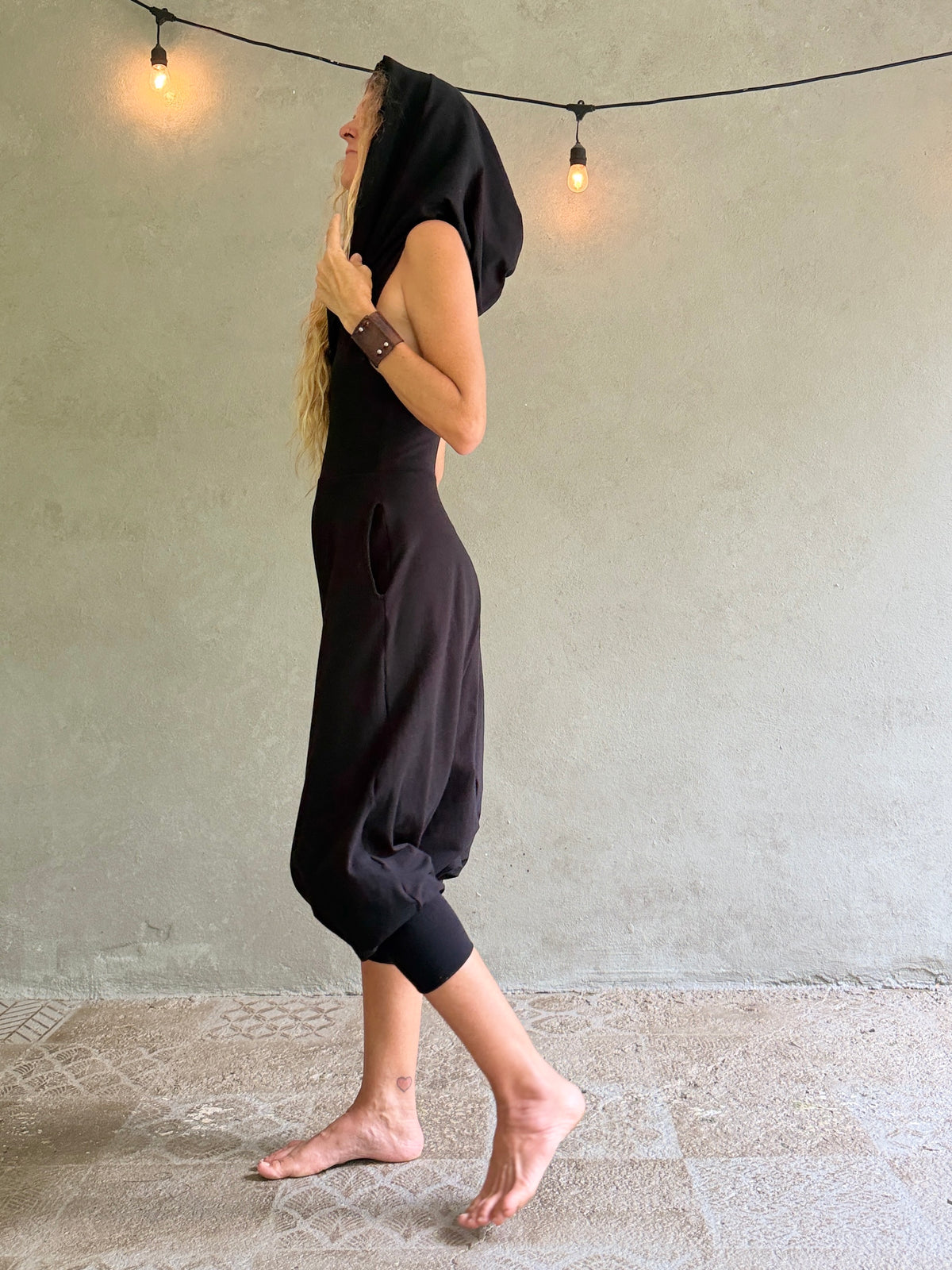 A woman wearing a loose black hooded jumpsuit or romper walks barefoot against a textured gray wall with hanging string lights.