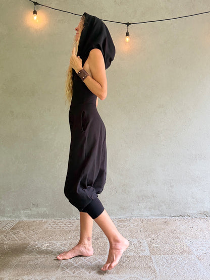 A woman wearing a loose black hooded jumpsuit or romper walks barefoot against a textured gray wall with hanging string lights.