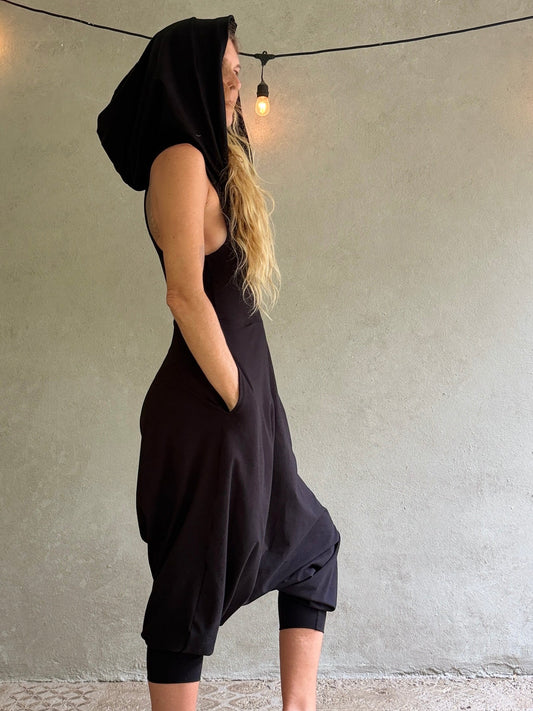 A woman wearing a black hooded sleeveless Aura jumpsuit with side pockets, standing in profile against a textured gray wall with a hanging light bulb.