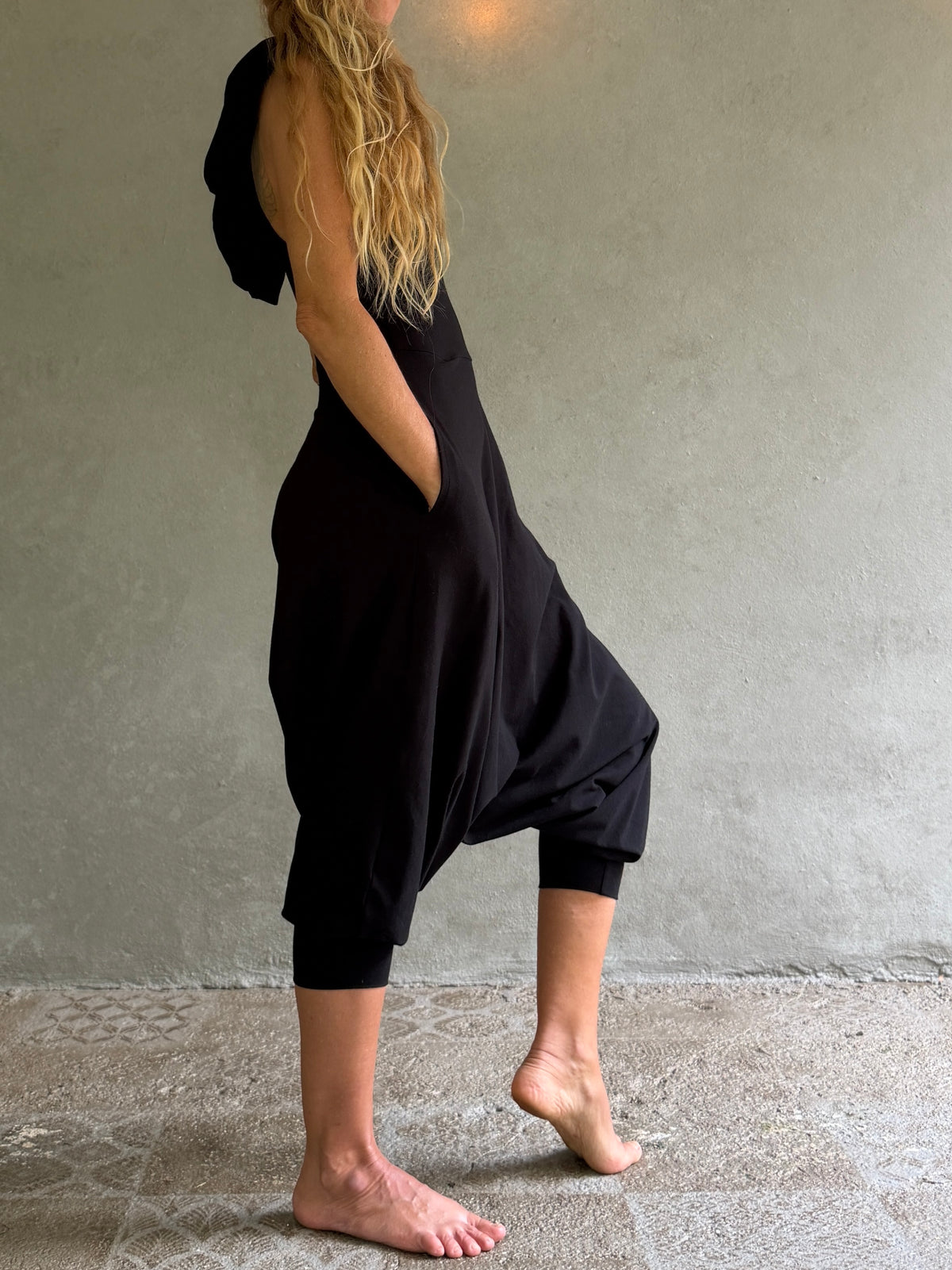A woman wearing a loose, black, one-shoulder harem-style jumpsuit with side pockets, standing barefoot against a textured gray concrete wall.