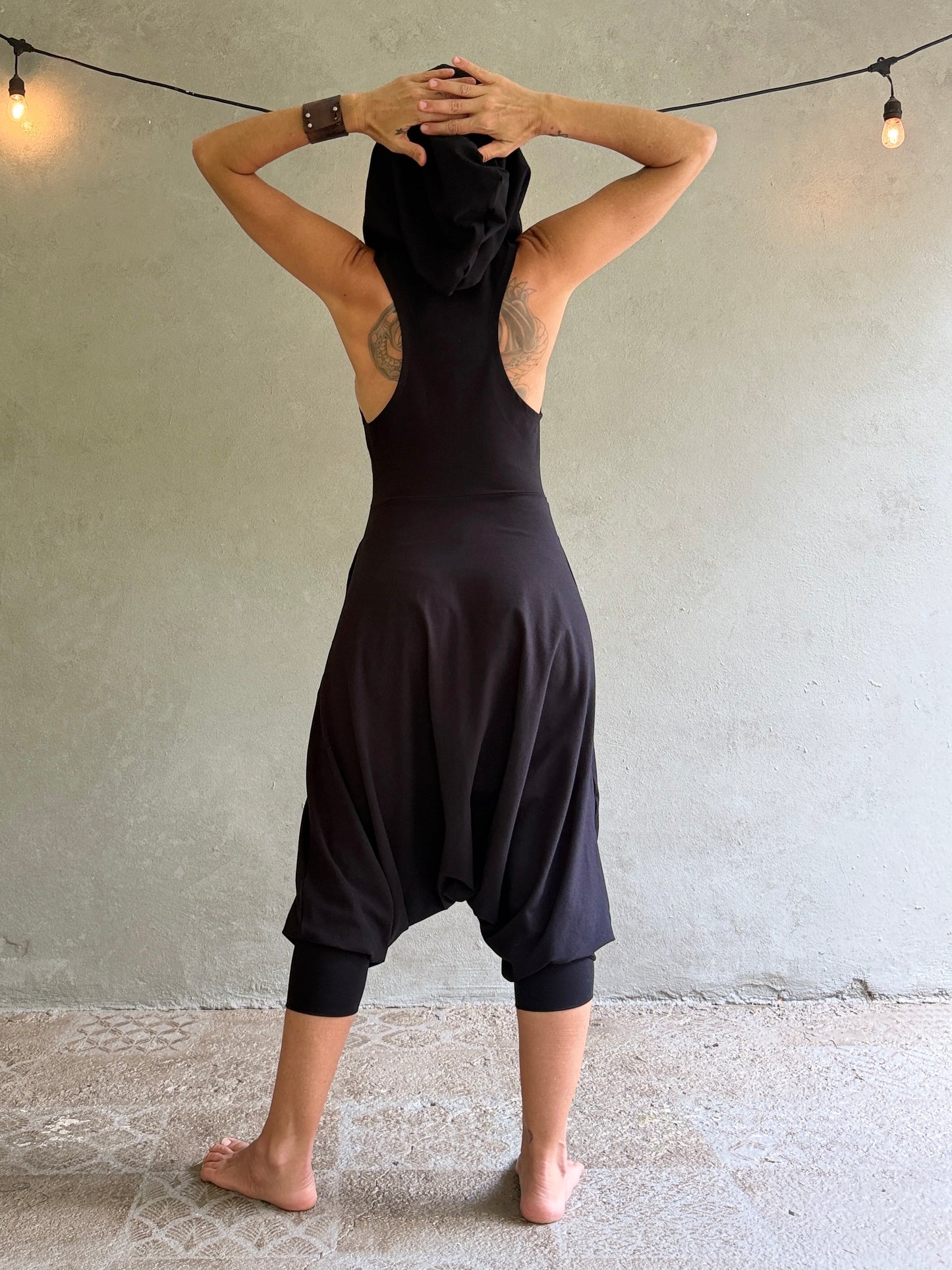 A woman stands with her back to the camera wearing a loose black harem-style jumpsuit with a high neck and cap sleeves, posed against a textured concrete wall with hanging string lights.