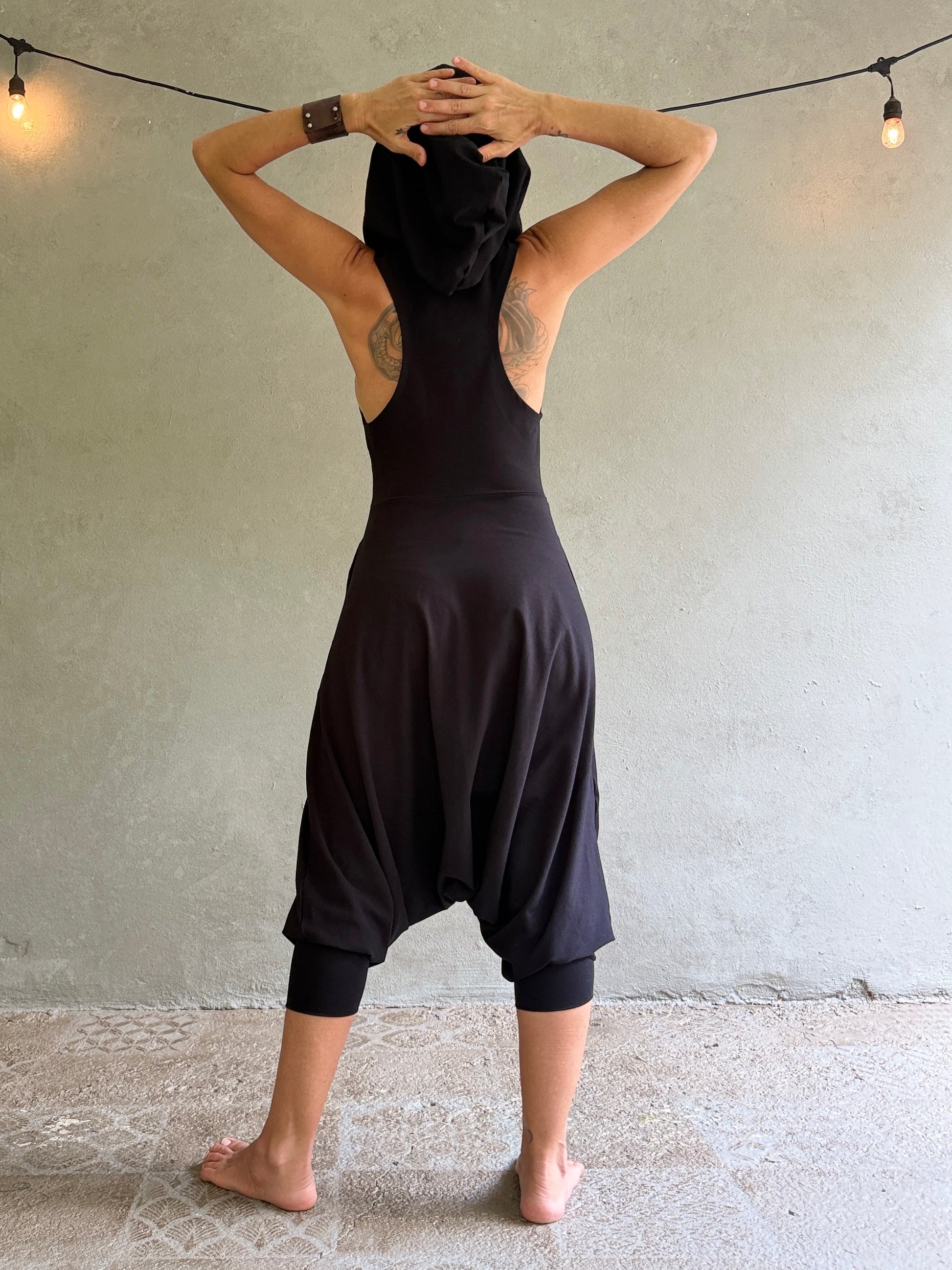 A woman stands with her back to the camera wearing a loose black harem-style jumpsuit with a high neck and cap sleeves, posed against a textured concrete wall with hanging string lights.
