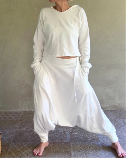 A woman wearing a white two-piece loungewear set consisting of a cropped long-sleeve top and matching high-waisted baggy pants, standing barefoot against a textured beige wall.