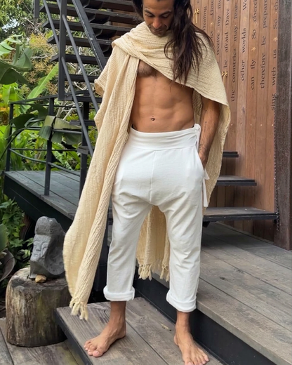 Man with long hair wearing a beige shawl and white glorka TOM pants standing on wooden steps.
