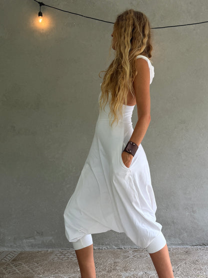 A woman wearing a white sleeveless harem-style Aura jumpsuit with side pockets, shown from the back against a textured gray wall with a hanging light bulb.