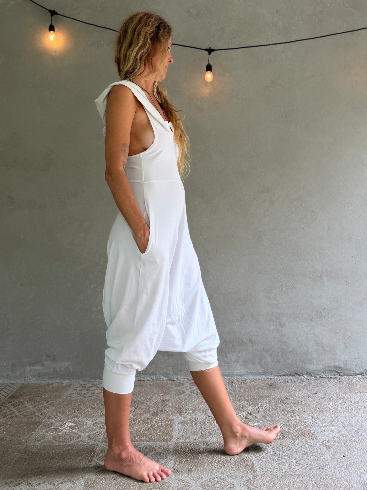 A woman wearing a white sleeveless harem-style Aura jumpsuit with side pockets, walking barefoot against a textured gray wall with hanging string lights.