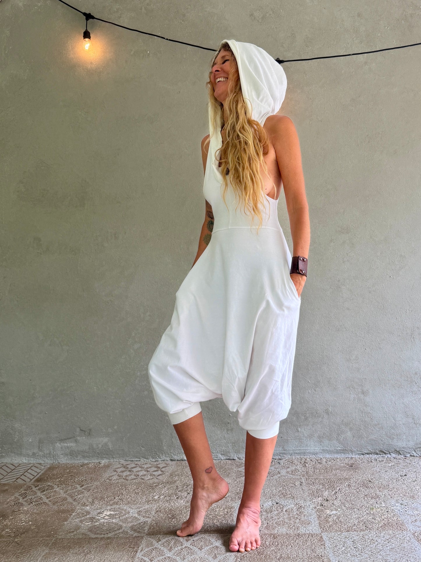 A woman wearing a white hooded sleeveless Aura jumpsuit with voluminous harem-style legs, standing barefoot against a textured gray wall with a hanging string light.