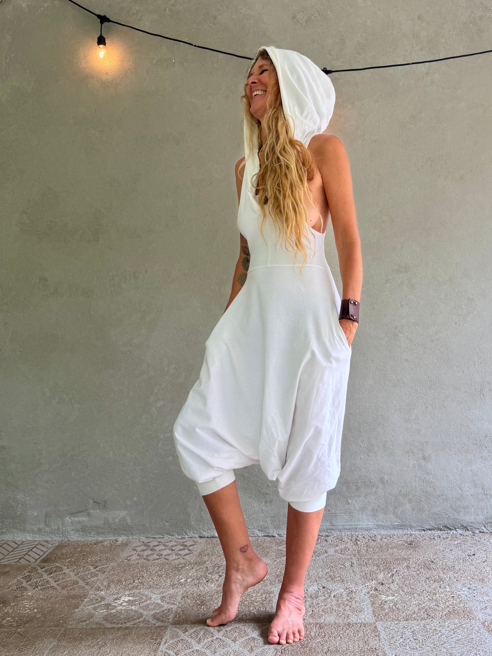 A woman wearing a white hooded sleeveless Aura jumpsuit with voluminous harem-style legs, standing barefoot against a textured gray wall with a hanging string light.
