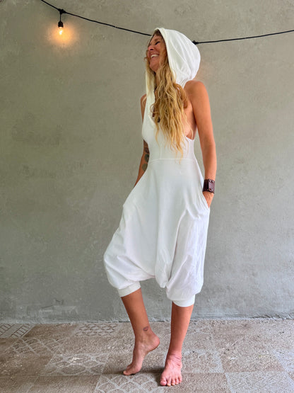 A woman wearing a white hooded sleeveless Aura jumpsuit with voluminous harem-style legs, standing barefoot against a textured gray wall with a hanging string light.
