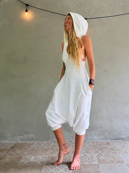 A woman wearing a white hooded sleeveless Aura jumpsuit with voluminous harem-style legs, standing barefoot against a textured gray wall with a hanging string light.