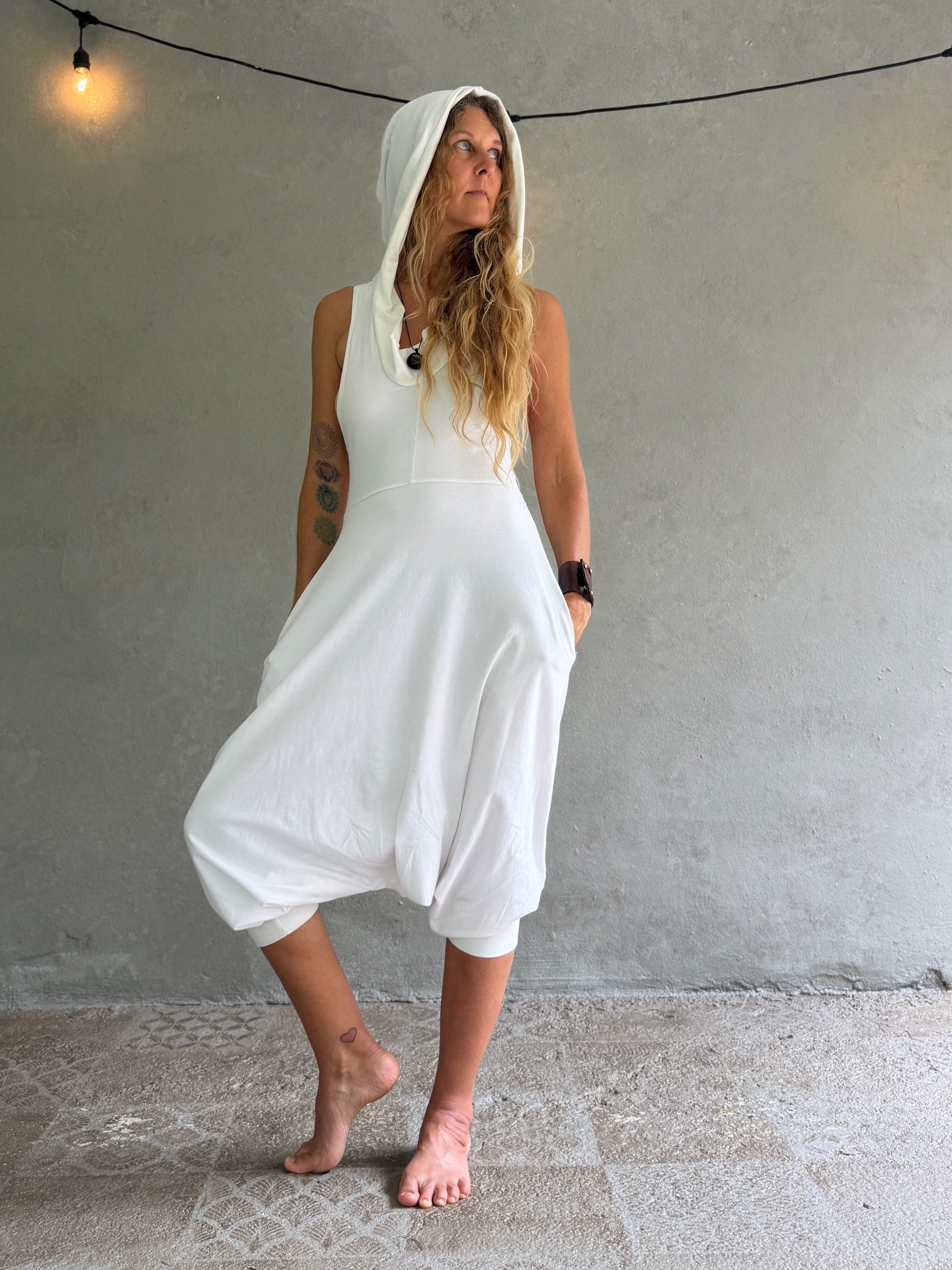 A woman wearing a white hooded sleeveless jumpsuit with large balloon-style legs, standing barefoot against a textured gray wall.