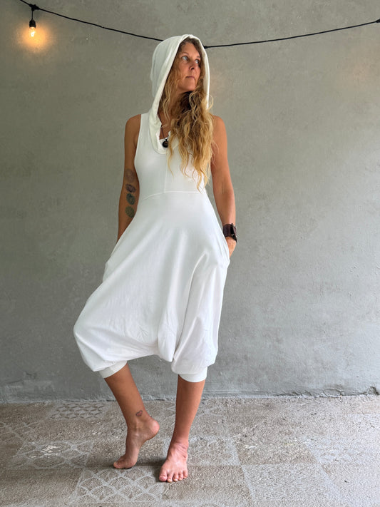 A woman wearing a white hooded sleeveless jumpsuit with large balloon-style legs, standing barefoot against a textured gray wall.