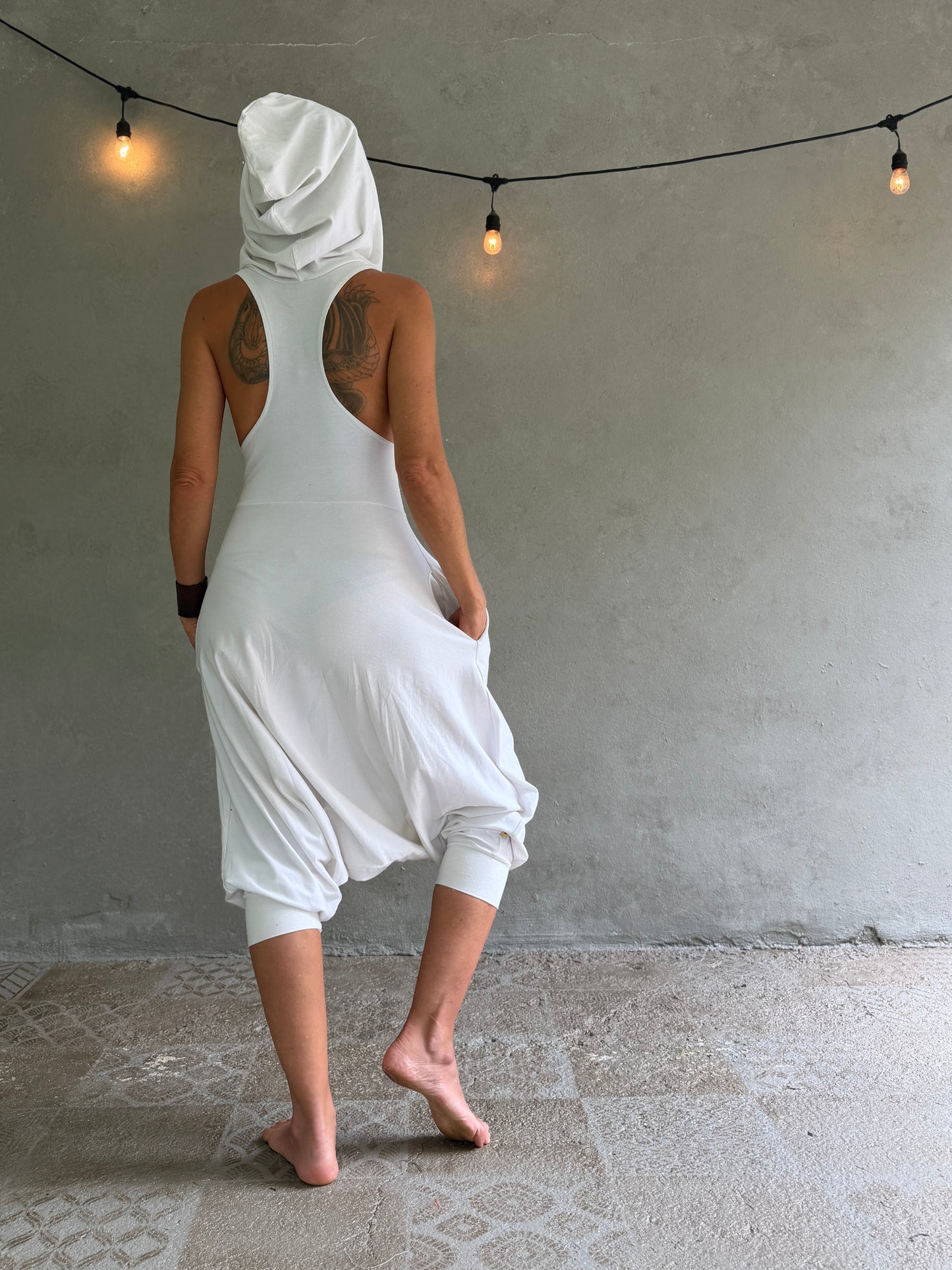 A woman stands with her back to the camera wearing a loose white hooded dress with a wide back and cuffed ankles, posed against a textured gray wall with hanging string lights.